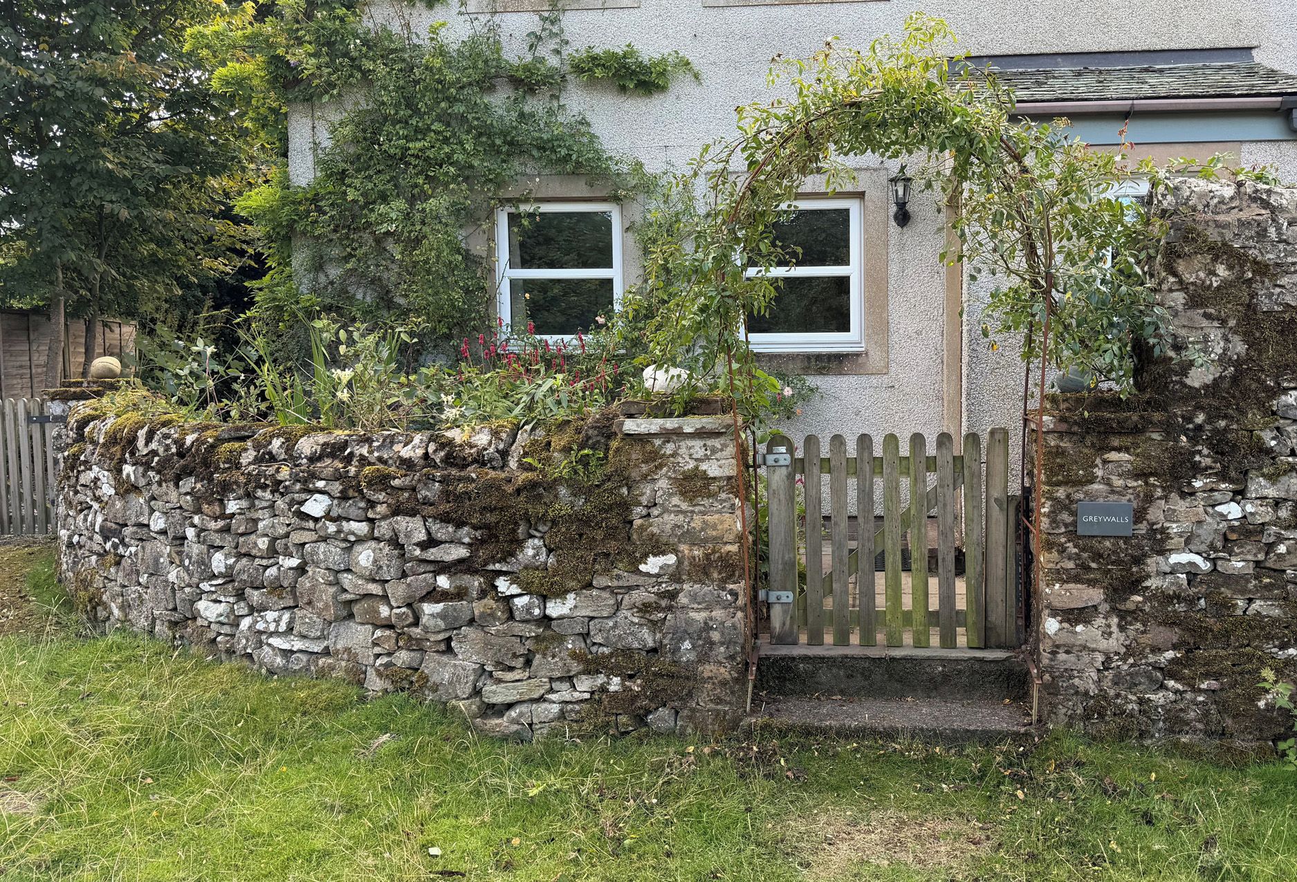 A stone wall with a wooden gate, a house with a window, and a sign reading 'GREYWALLS'.