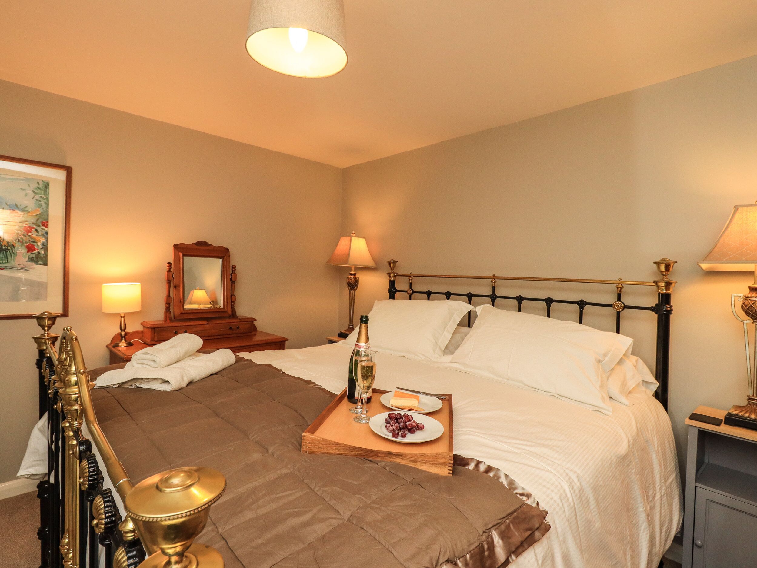 A neatly made bed with white linens, a bedside table with a lamp, a tray with a bottle of champagne, and a framed picture on the wall.