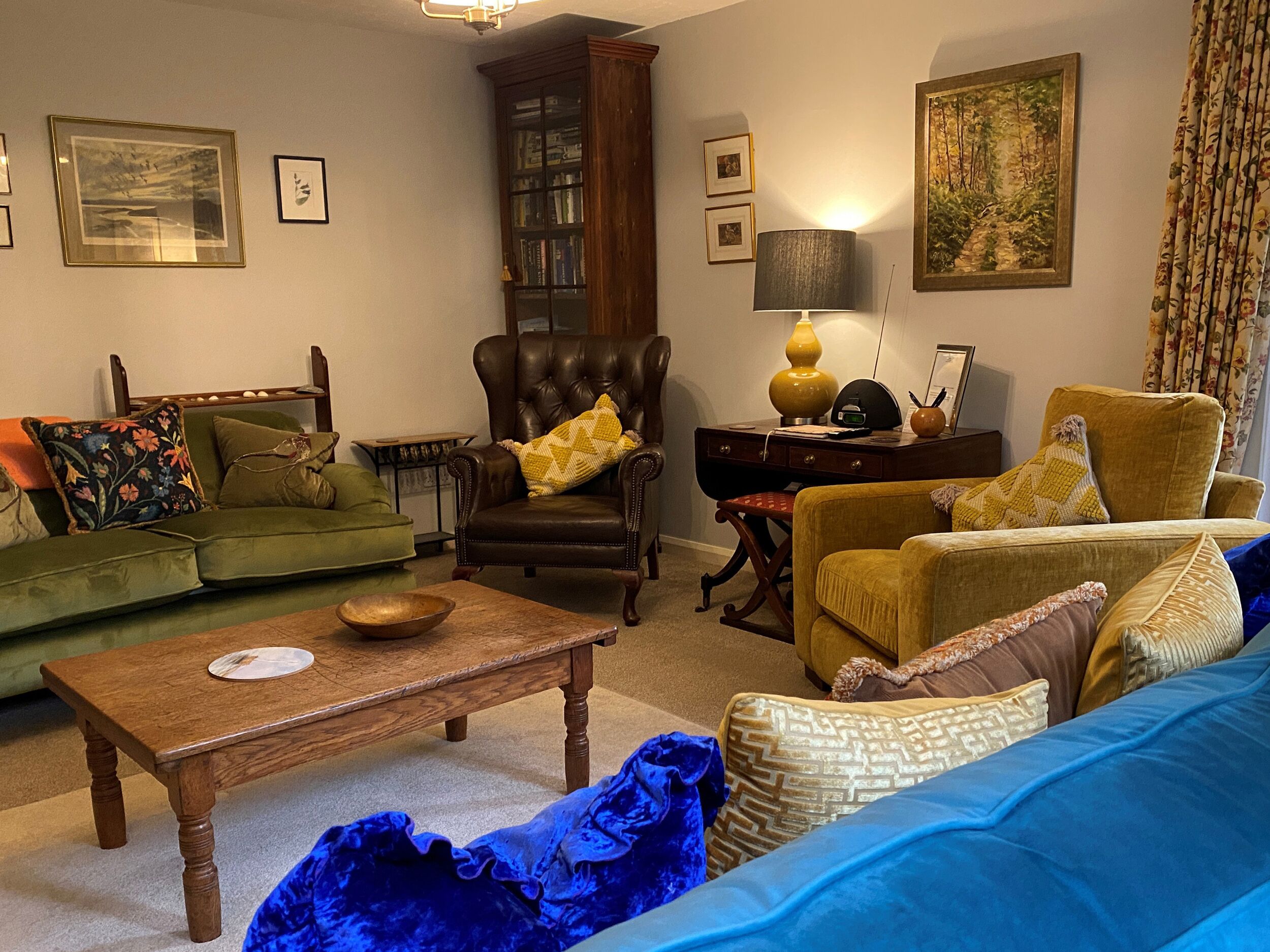 Een woonkamer met een groene bank, een bruine leren fauteuil, een houten salontafel en een blauwe bank. Er hangen fotolijstjes aan de muren en staat een lamp op een bijzettafeltje.