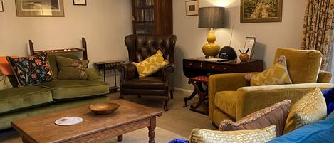 Een woonkamer met een groene bank, een bruine leren fauteuil, een houten salontafel en een blauwe bank. Er hangen fotolijstjes aan de muren en staat een lamp op een bijzettafeltje.