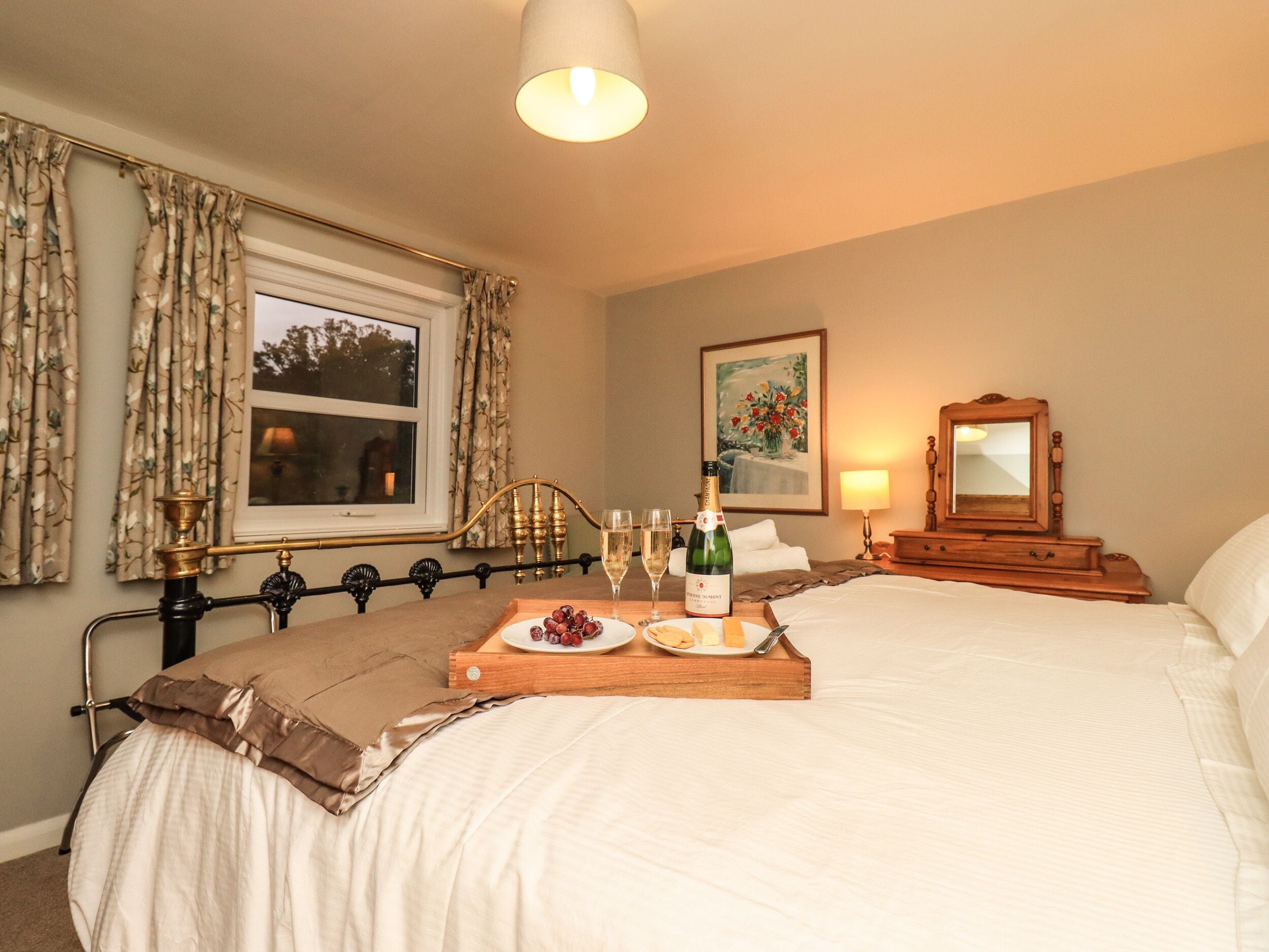 A hotel room with a bed, a bedside table with a bottle of champagne, a tray with grapes and cheese, and a painting on the wall.