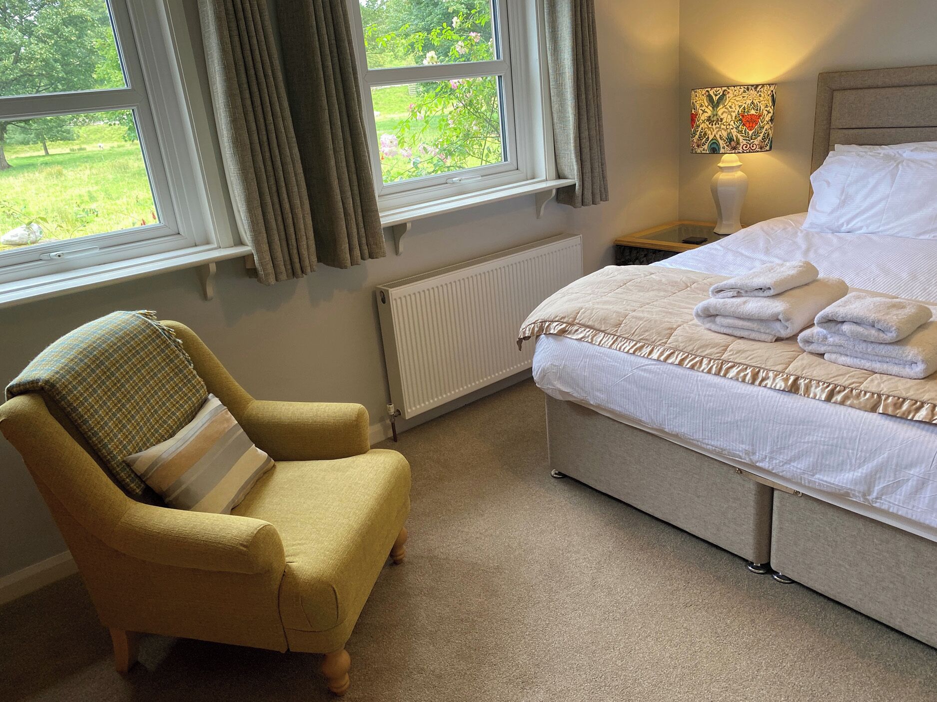 A bedroom with a bed, a chair, a window with curtains, a radiator, and a wall-mounted lamp.