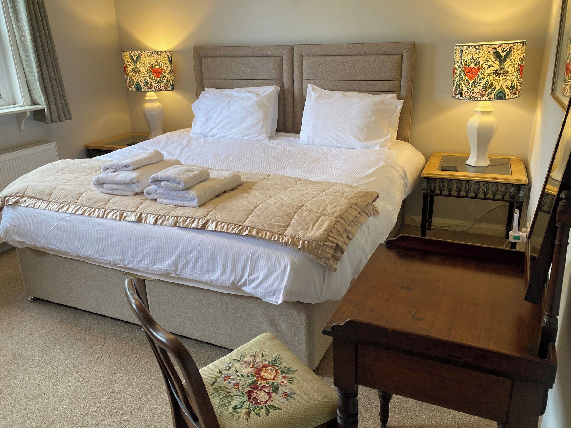 A neatly made bed with white linens, two bedside lamps with floral patterns, a wooden nightstand, and a chair with a floral cushion.