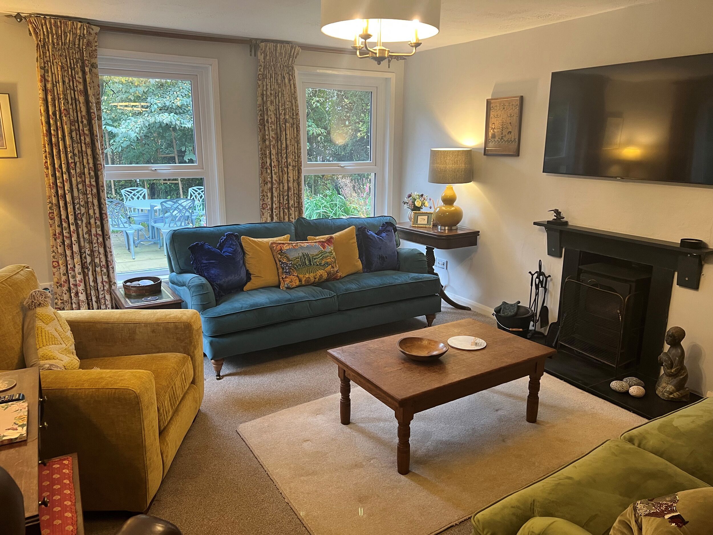 A living room with a blue sofa, yellow armchair, and green armchair. There is a wooden coffee table, a fireplace, and a television mounted on the wall.