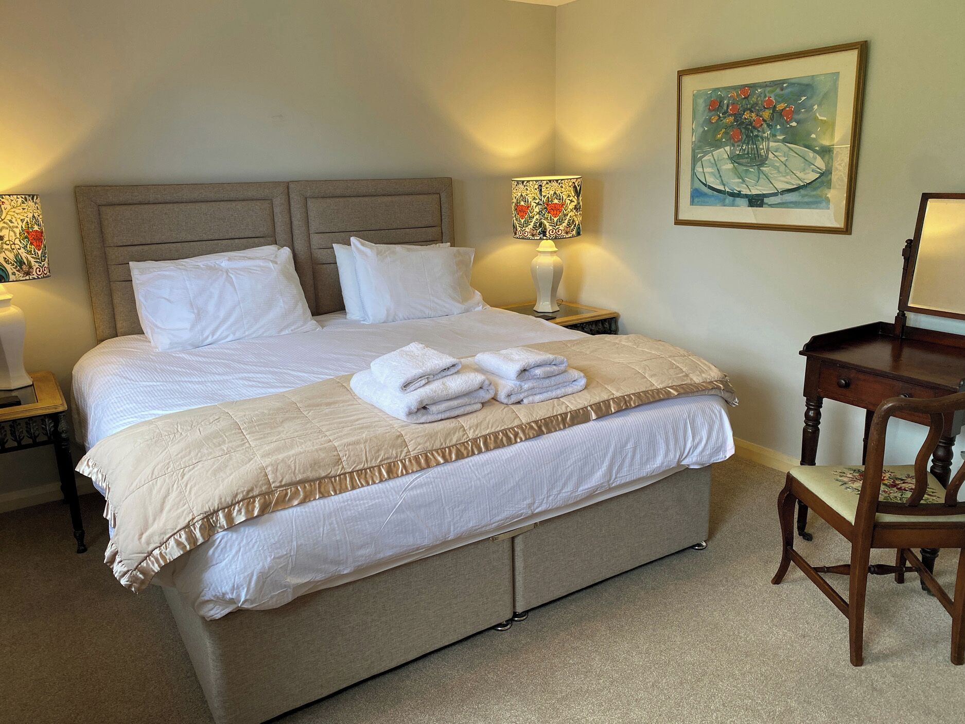 A neatly made bed with white linens and a beige quilt, two bedside lamps, a wooden nightstand, and a chair with a floral cushion.