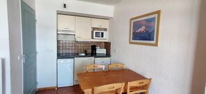 Eine kleine Küche mit weißen Schränken, einer Mikrowelle und einem Toaster. Ein Holztisch bietet Platz für vier Stühle. An der Wand hängt ein gerahmtes Bild einer Berglandschaft.