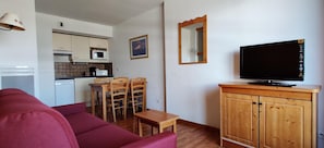 Ein Wohnzimmer mit rotem Sofa, einem Holzespeisetisch und Stühlen, einem Fernseher auf einer Holzkommode und einer Küche im Hintergrund.