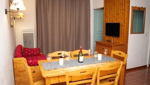 A cozy room with a wooden dining table set for four, a red sofa, a television, and a wooden cabinet. - 300m from ski slopes, Ground floor, swimming pool, sauna, balcony, parking, tv, ski locker, 30m² (Le Dévoluy)