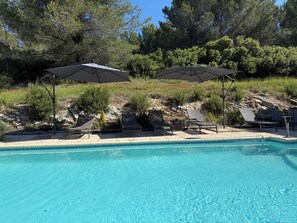 A swimming pool with lounge chairs and umbrellas, surrounded by trees and a hilly landscape.
