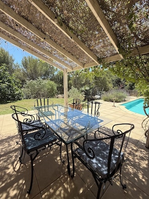 Une terrasse extérieure avec une table en verre et des chaises en métal, entourée d’une pergola illuminée de guirlandes lumineuses.