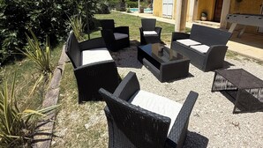 En utomhusplats med sittmöbler i rotting och ett stort parasoll, omgiven av en trädgård och en byggnad med ett tak täckt av tegelpannor.