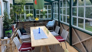 Ein überdachter Patio-Bereich mit Holztisch, weißen Plastikstühlen und Blumentöpfen.