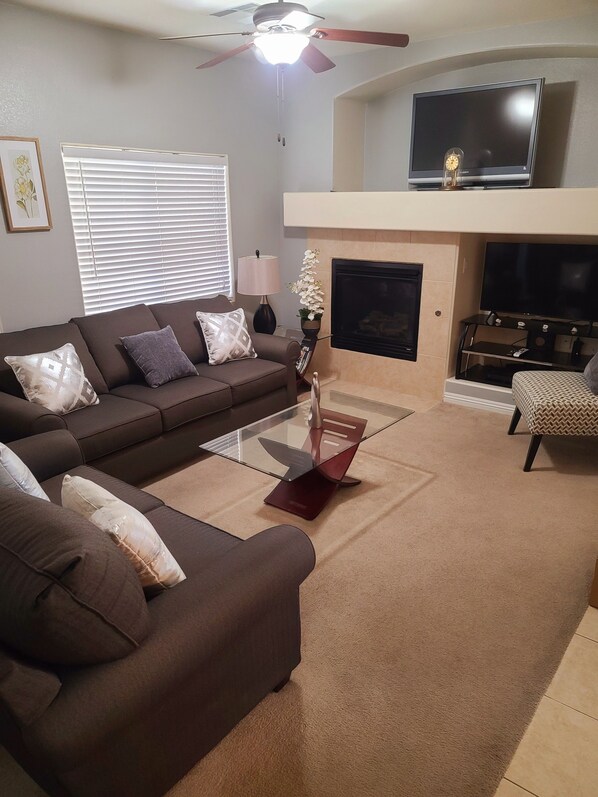 A living room with a brown sofa, a glass coffee table, a fireplace, a television, and a ceiling fan.