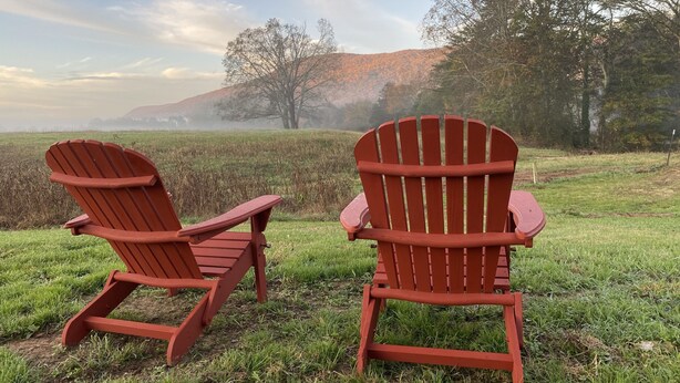 Two red Adirondack chairs facing each other in a grassy field with a misty background of trees and hills.