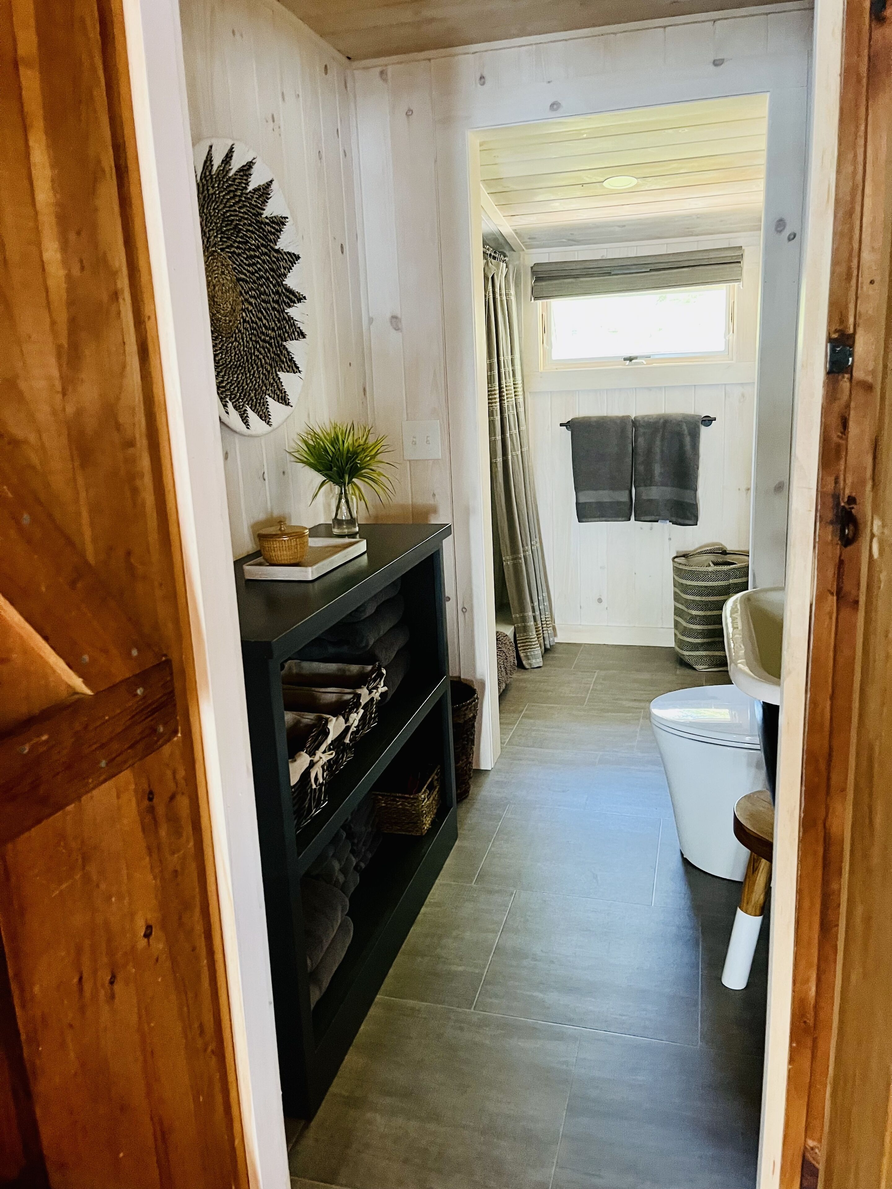 Un corridoio stretto con una porta in legno a sinistra, una mensola nera con oggetti decorativi, un water bianco e una finestra con tende.