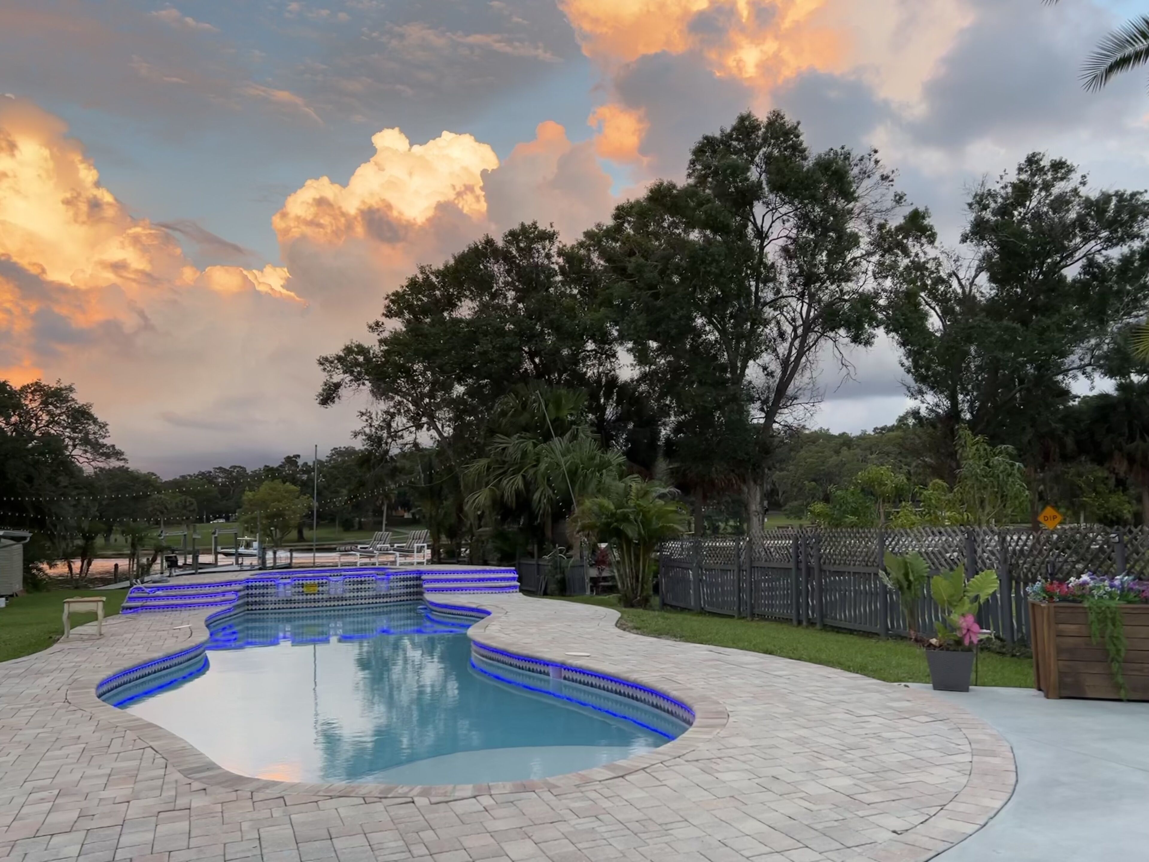 A swimming pool with blue LED lights, surrounded by a brick patio, trees, and a wooden fence.