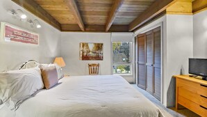 Ein Schlafzimmer mit einem großen Bett, einem hölzernen Kopfteil, einem Sessel, einem Schreibtisch mit Fernseher und einem Fenster mit Rollläden.