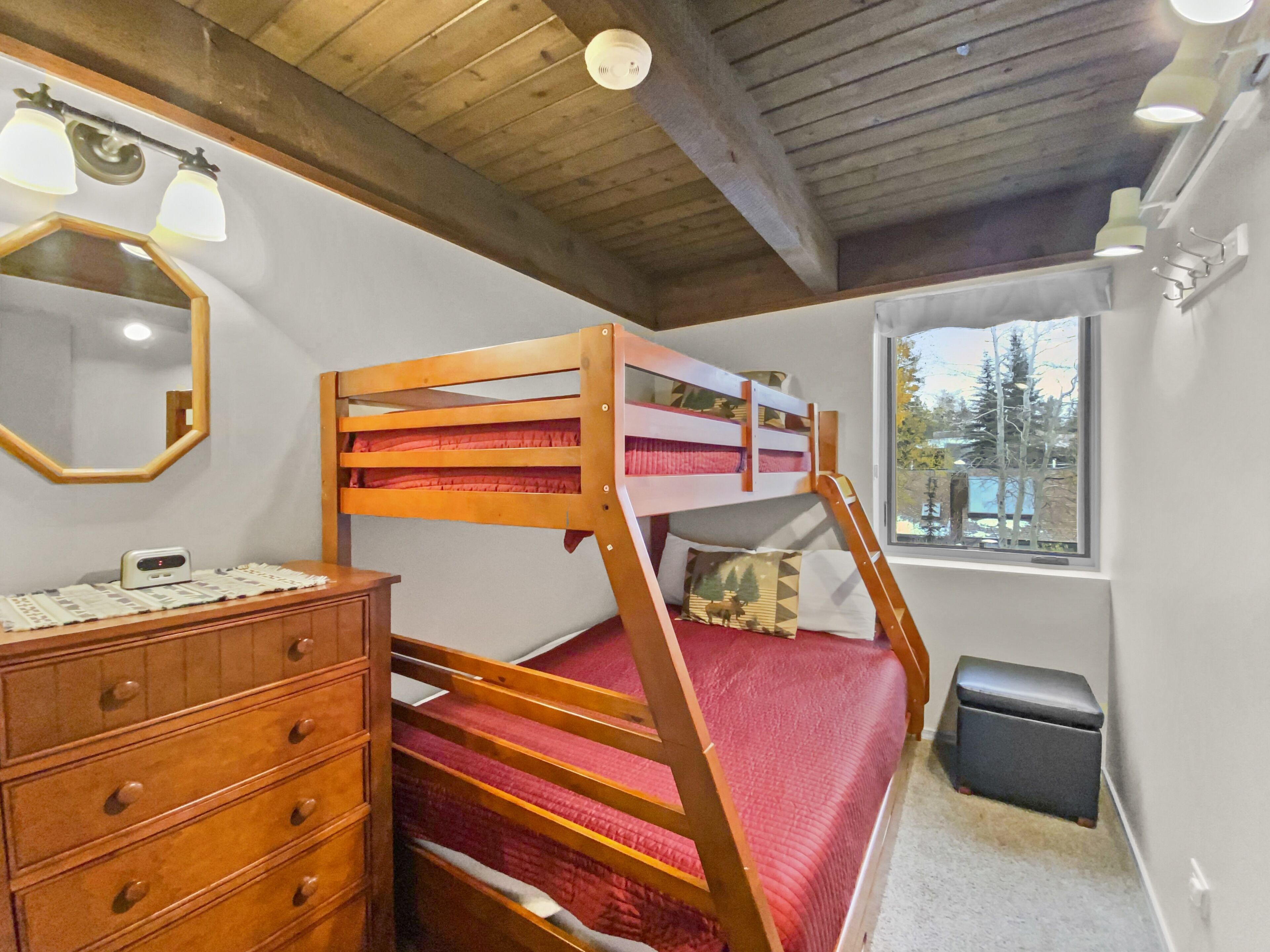 A bunk bed with a wooden frame and red bedding, a dresser with drawers, a mirror, and a window with a view of trees.