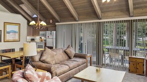 Ein gemütliches Wohnzimmer mit einer braunen Couch, einem gemusterten Sessel und einem Holztisch. Im Hintergrund ist eine Küche mit Mikrowelle und Kühlschrank zu sehen.