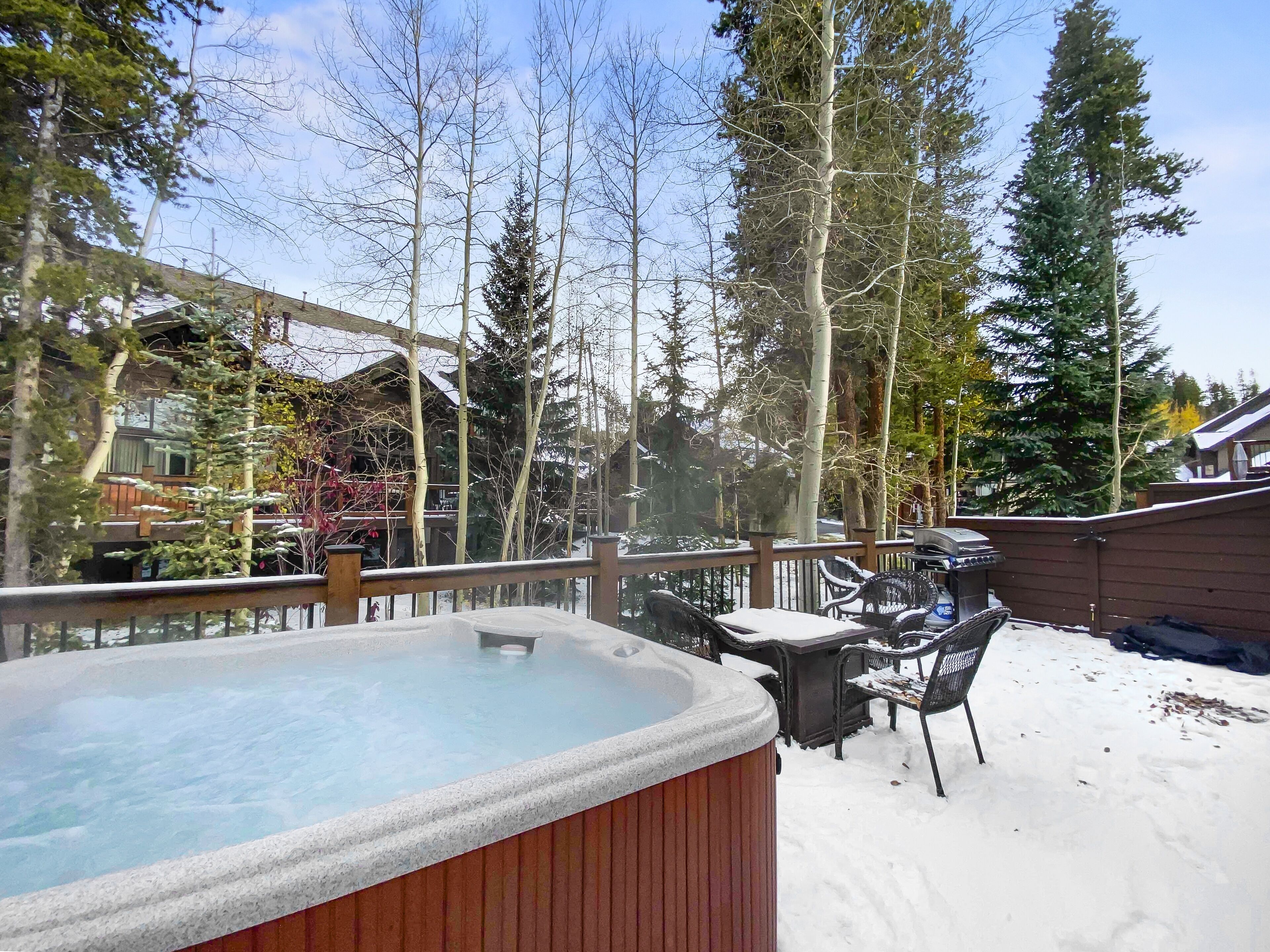 A hot tub surrounded by snow-covered ground, wooden deck, and outdoor furniture.