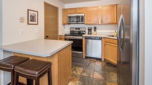 Una cocina con armarios de madera, un refrigerador de acero inoxidable, un microondas, un horno y un lavavajillas. Hay dos taburetes en la barra.