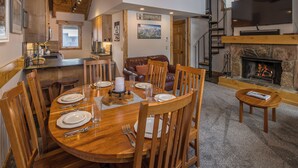 A dining area with a wooden table set for four, surrounded by chairs, a fireplace, and a television mounted above it. - Big Sky Town Center condo w/fireplace & community pool/hot tub/sauna - The Grateful Getaway (Big Sky)
