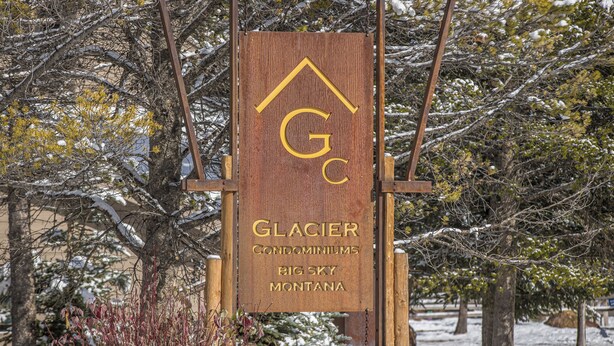 A wooden sign with the text "Glacier Condominiums Big Sky Montana" and a logo. - Big Sky Town Center condo w/fireplace & community pool/hot tub/sauna - The Grateful Getaway (Big Sky)