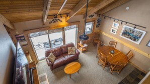 A cozy living room with a brown leather sofa, a wooden coffee table, and a dining table with chairs. There are framed pictures on the walls and a large window providing a view outside. - Big Sky Town Center condo w/fireplace & community pool/hot tub/sauna - The Grateful Getaway (Big Sky)