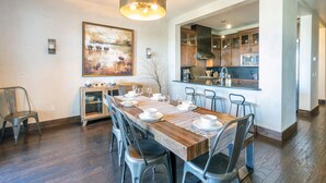 A spacious dining area with a long wooden table set for six, surrounded by modern chairs. A large abstract painting adorns the wall above the table. - CASSIDY'S HIDEOUT- Beautiful Condo with Amazing San Sophia Ridge Views (Mountain Village)