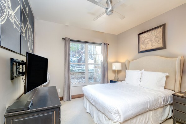 A bedroom with a bed, a TV, a desk, a window with curtains, a ceiling fan, and a framed map on the wall.