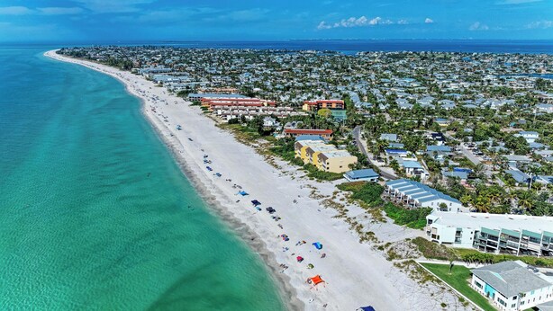 An aerial view of a coastal town with a long sandy beach, several buildings, and a clear blue sky. - Osprey in Blue Lagoon Unit 6 - Condo with Pool - By Island Real Estate Vacations (Holmes Beach)