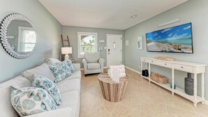 Ein Wohnzimmer mit einem weißen Sofa, blauen Kissen mit Muster, einem runden Couchtisch, einem weißen Konsolentisch mit Fernseher und einem Gemälde mit Strandmotiv.
