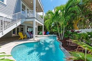 A swimming pool with a curved design, surrounded by a wooden deck and a covered patio area with chairs.