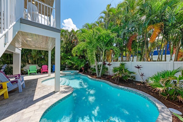 A swimming pool with a covered area, surrounded by colorful Adirondack chairs and lush tropical plants.