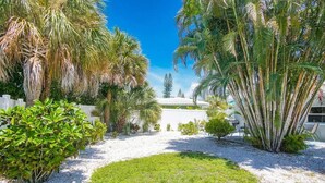 A tropical garden with palm trees, a white fence, and a building with a green roof. - Beach Life by the Gulf- Charming & Fully Equipped! (Redington Beach)