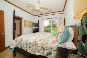 A bedroom with a bed, a ceiling fan, a television, and a window with a view of greenery.