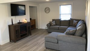 Una sala de estar con un sofá seccional gris, un mueble de entretenimiento de madera, un televisor de pantalla plana y un reloj en la pared.