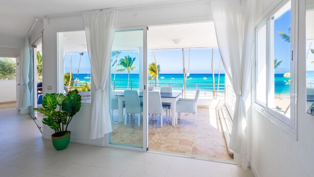 A room with a view of the beach, a dining area with chairs and a table, and a large potted plant. - Ocean View Luxury Apartment Directly on the Los Corales Beach (Punta Cana)