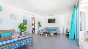 A modern living room with a sofa, coffee table, and plants. There is a TV mounted on the wall and a window with curtains. - Punta Cana Beachfront Penthouse - Ocean View, BBQ, Terrace, 4BR (Punta Cana)