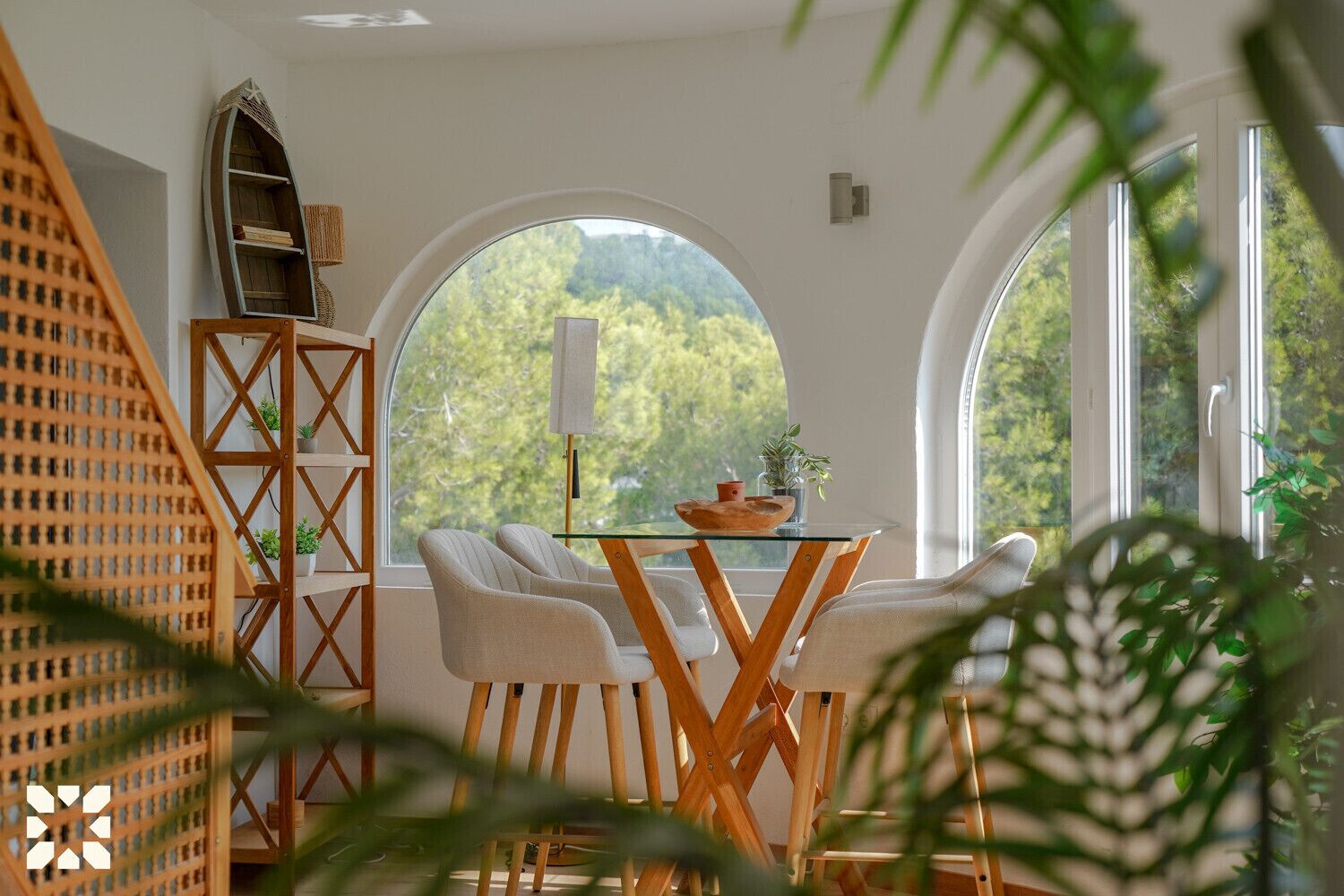 A room with a round table, two chairs, a bookshelf, and a view of trees through a large window.