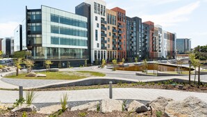 Un immeuble moderne à plusieurs étages, dont la façade est un mélange de verre et de briques, avec une cour aménagée dotée de sentiers et d’un petit élément décoratif aquatique.
