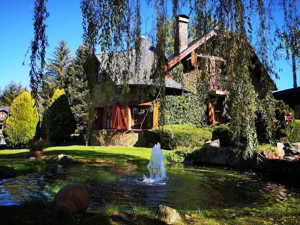 Una casa rústica con chimenea de piedra, un estanque con fuente y abundante vegetación.