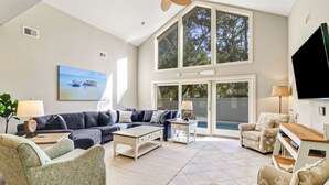 A spacious living room with a large sectional sofa, a coffee table, and a patterned armchair. There is a painting on the wall, a television, and a large window with a view of trees. - 3rd Row Ocean - Dog Friendly Sea Pines Home w/ FREE Pool Heat Seasonally! (Hilton Head Island)