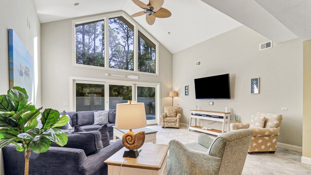 A spacious living room with a high ceiling, large windows, a ceiling fan, a flat-screen TV, and a comfortable seating area with a sofa and armchairs. - 3rd Row Ocean - Dog Friendly Sea Pines Home w/ FREE Pool Heat Seasonally! (Hilton Head Island)