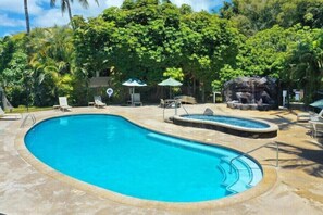 Une piscine avec un jacuzzi, entourée de chaises longues et de parasols, le tout dans un cadre verdoyant et luxuriant.