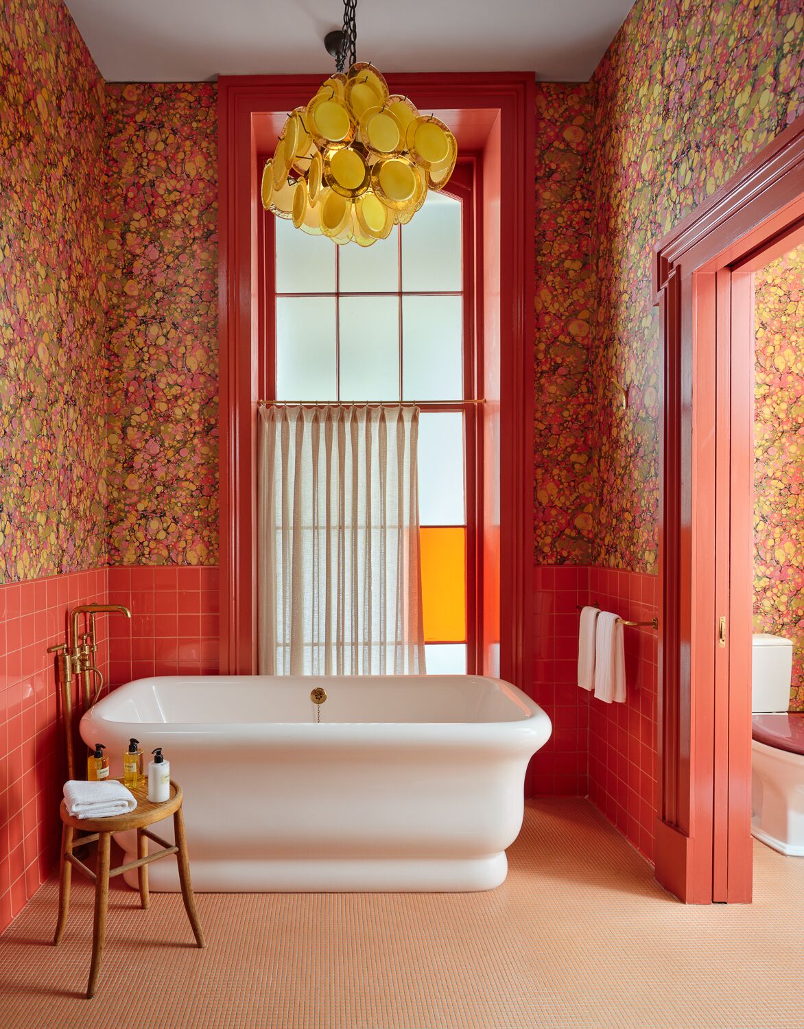 A bathroom with a freestanding bathtub, a wooden stool with toiletries, and a unique yellow chandelier.