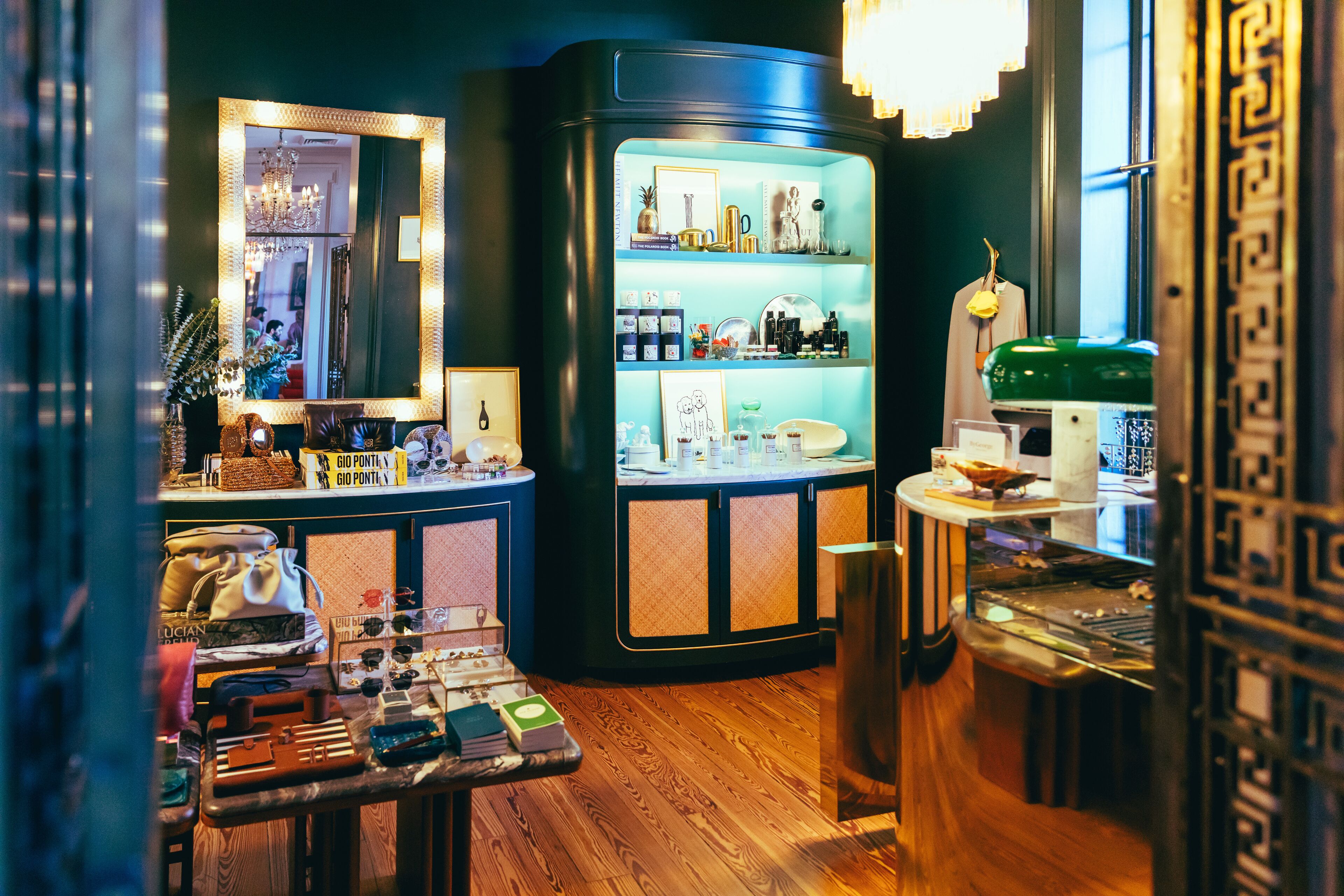 Interieur van een boetiekhotel met een houten vloer, een spiegel met gouden lijst, een vitrinekast met diverse objecten, en een groene koffer op een tafel.