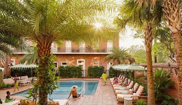 Zona de piscina con sillones de descanso y sombrillas, rodeada de vegetación y un edificio con balcón.