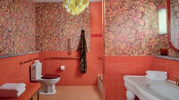 Une salle de bain avec un papier peint à motifs floraux, un lavabo blanc, des toilettes et un banc avec des serviettes.