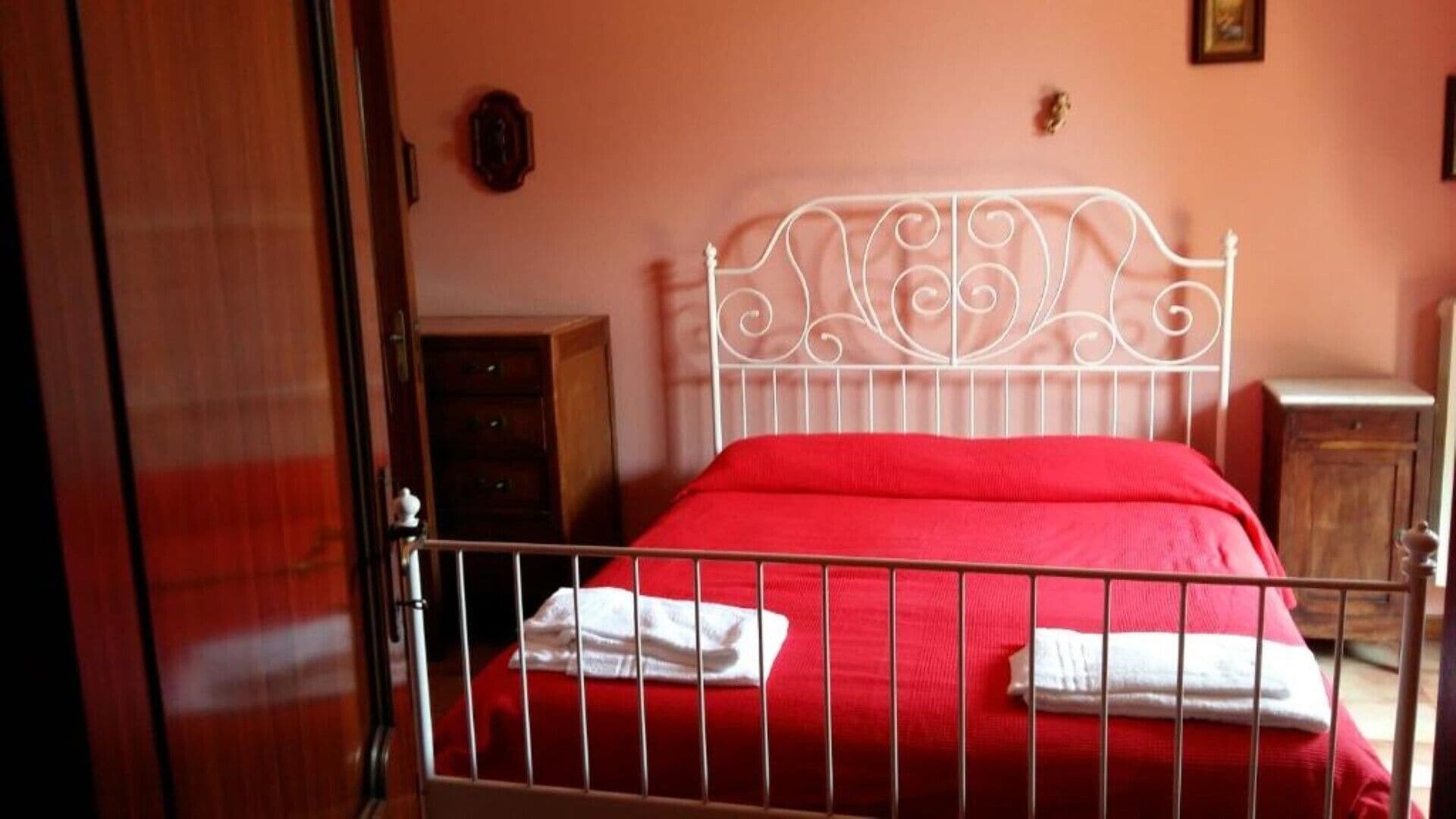 Una camera da letto con una struttura letto in metallo, una coperta rossa e asciugamani bianchi.